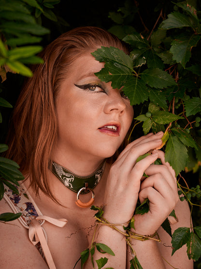Woman with red hair and dark makeup holding green leaves, surrounded by foliage wearing a bdsm snake collar