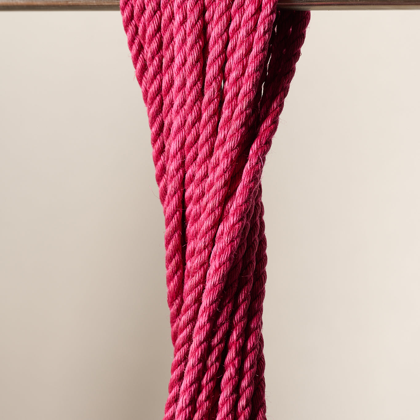 A close-up of a thick, 6mm, jute, twisted magenta pink rope draped over a horizontal metal rod against a plain light beige background.