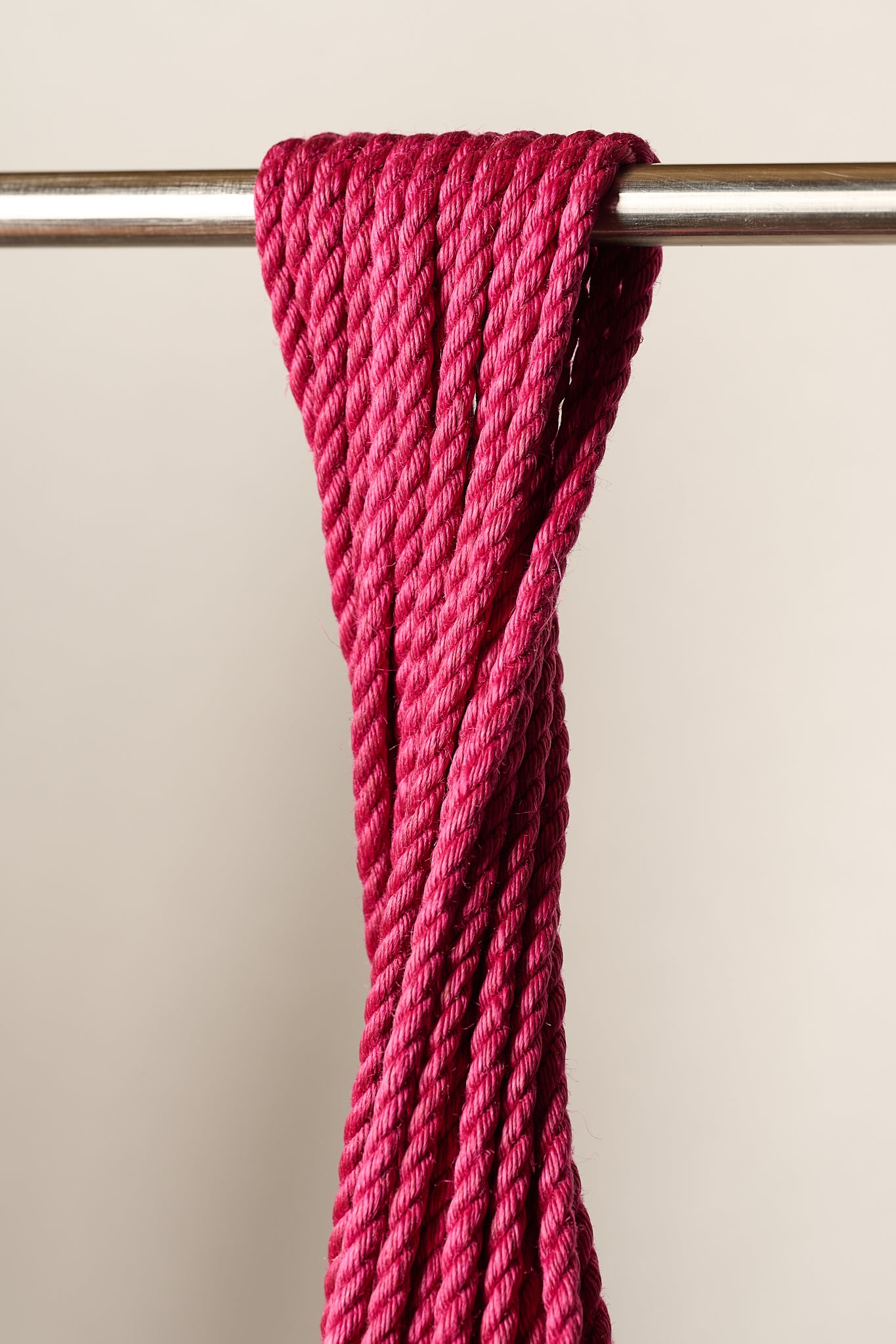 A close-up of a thick, 6mm, jute, twisted magenta pink rope draped over a horizontal metal rod against a plain light beige background.