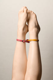 A pair of crossed legs with single strap connective ankle bracelets, one side pink with a silver buckle and other side  mustard yellow, shown against a neutral light gray background.