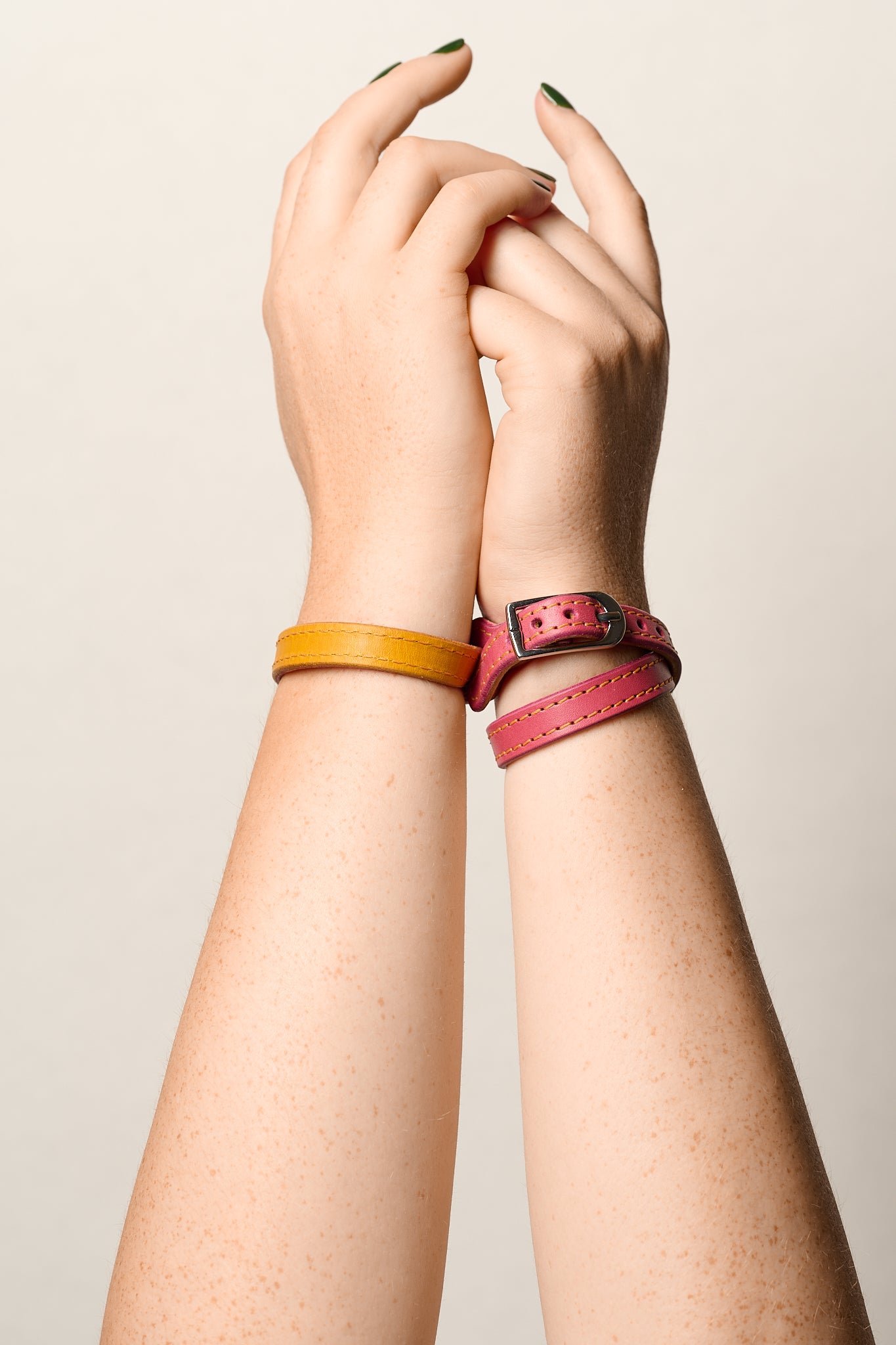 Two women's leather bracelets on wrists, one in mustard yellow and the other in pink with a silver buckle, shown on a model's arms against a light beige background.