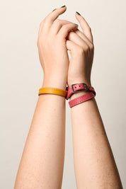 Two women's leather bracelets on wrists, one in mustard yellow and the other in pink with a silver buckle, shown on a model's arms against a light beige background.