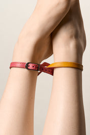 A close-up of a person's crossed legs wearing two slim leather ankle bracelets, one in pink with a silver buckle and one in mustard yellow.