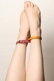 A pair of crossed bare feet and lower legs wearing two ankle bracelets: one pink leather strap with a silver buckle, and one mustard yellow leather bracelet, against a plain light beige background.