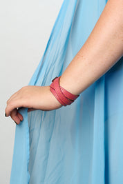 A close-up of a person's hand and wrist wearing a multi-strand pink leather wrap bracelet, with a light blue sheer dress draped in the background.