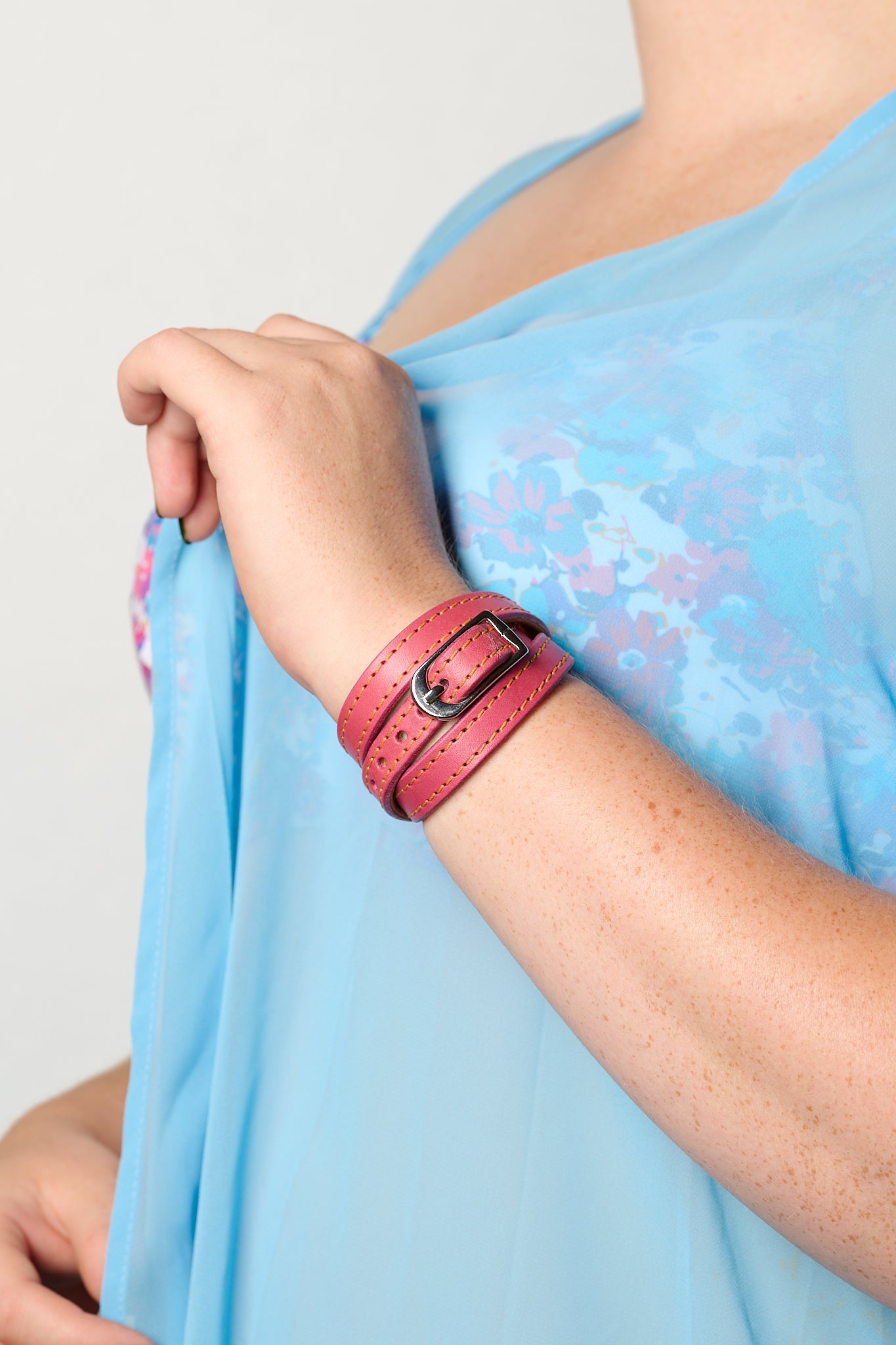 A close-up of a person wearing a pink leather triple-strand bracelet with a silver buckle, paired with a light blue floral printed top.
