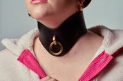 Person wearing a wide black choker with a gold ring and pink jacket on a gray background