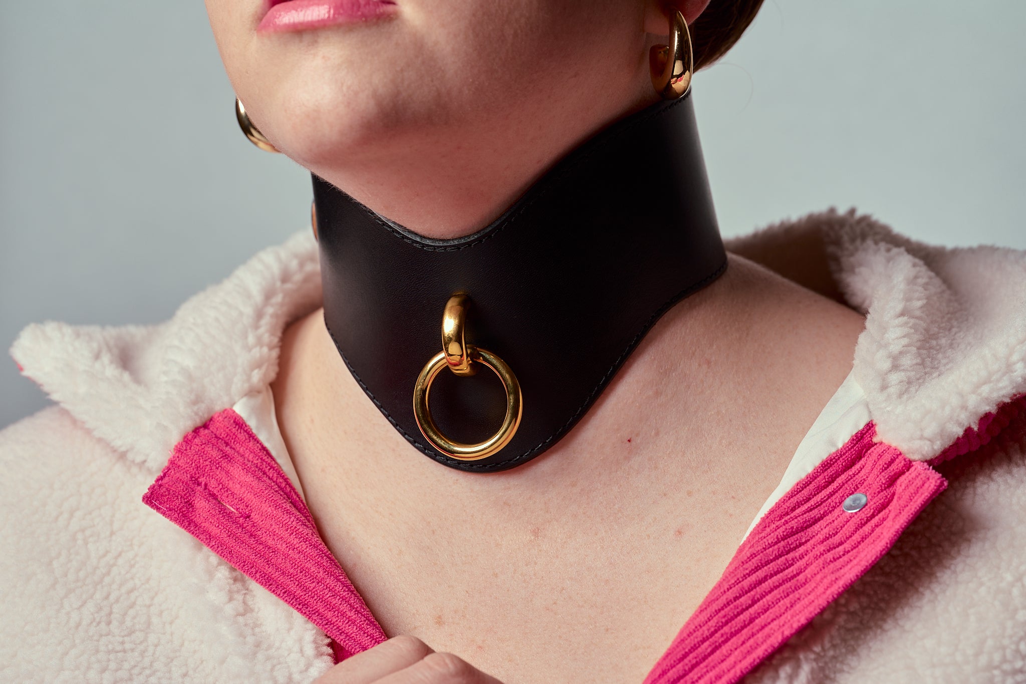 Person wearing a wide black choker with a gold ring and pink jacket on a gray background