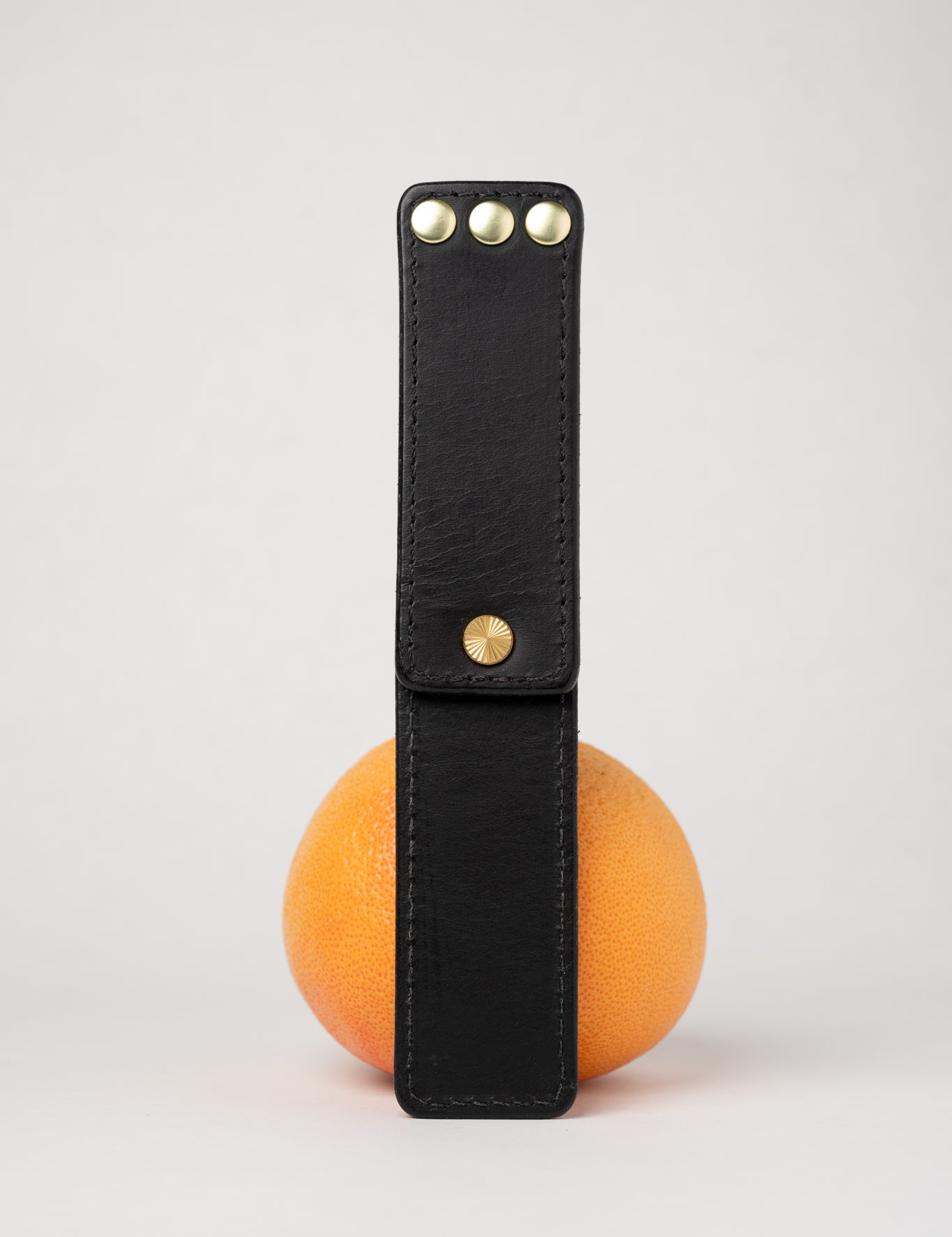 foldable black  leather travel size belt with rivets on white background with grapefruit behind.
