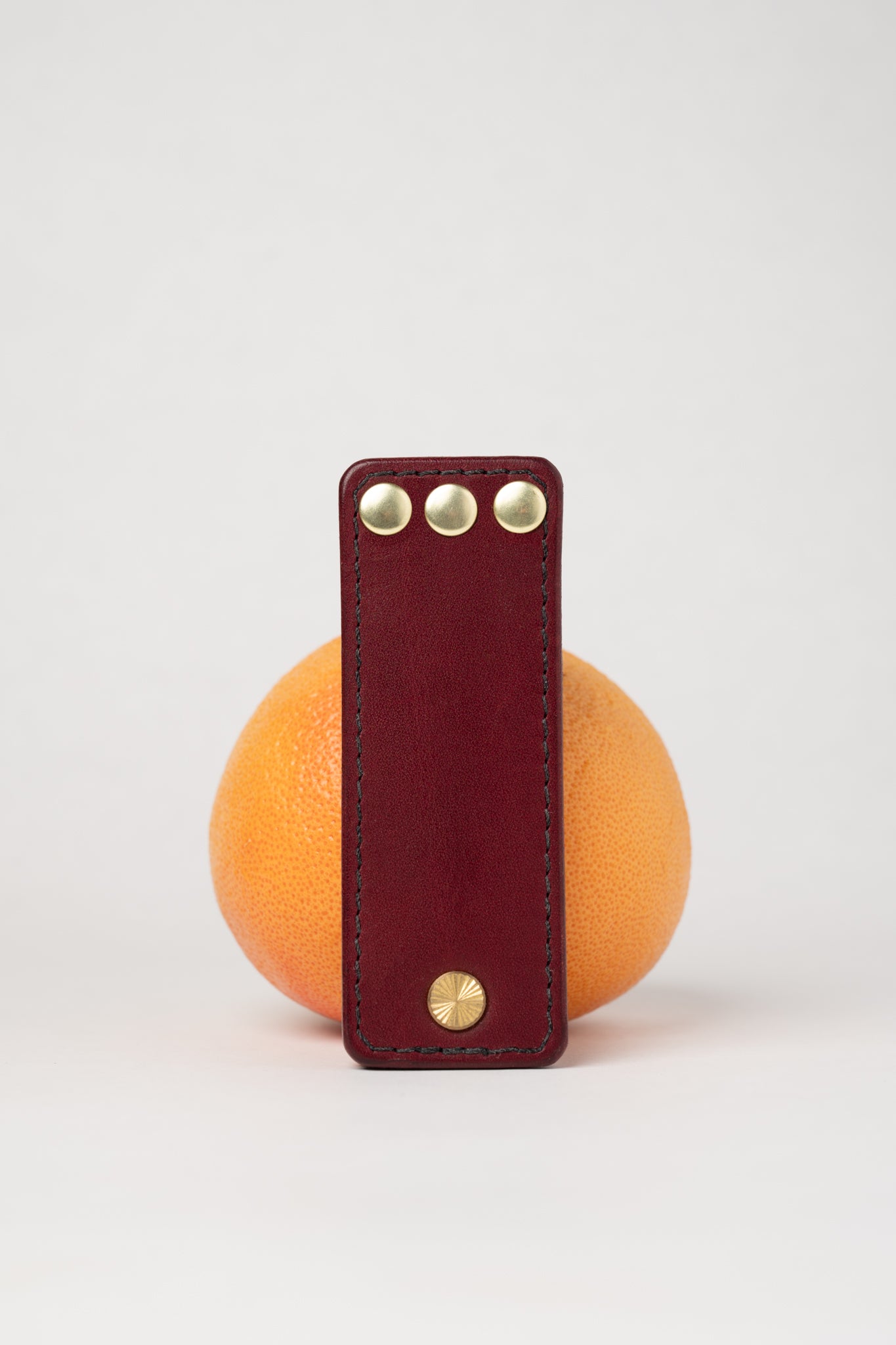foldable burgundy leather travel size belt with rivets on white background with grapefruit behind.