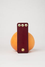 foldable burgundy leather travel size belt with rivets on white background with grapefruit behind.