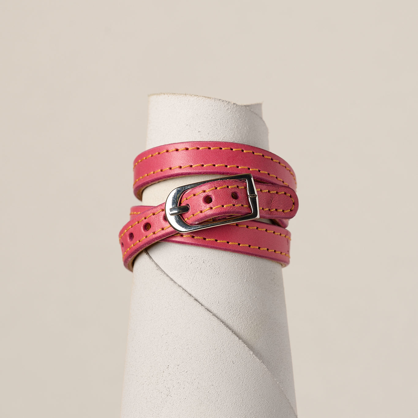 A pink leather belt with a silver-toned metal buckle, wrapped around a white cylindrical display stand against a light beige background.