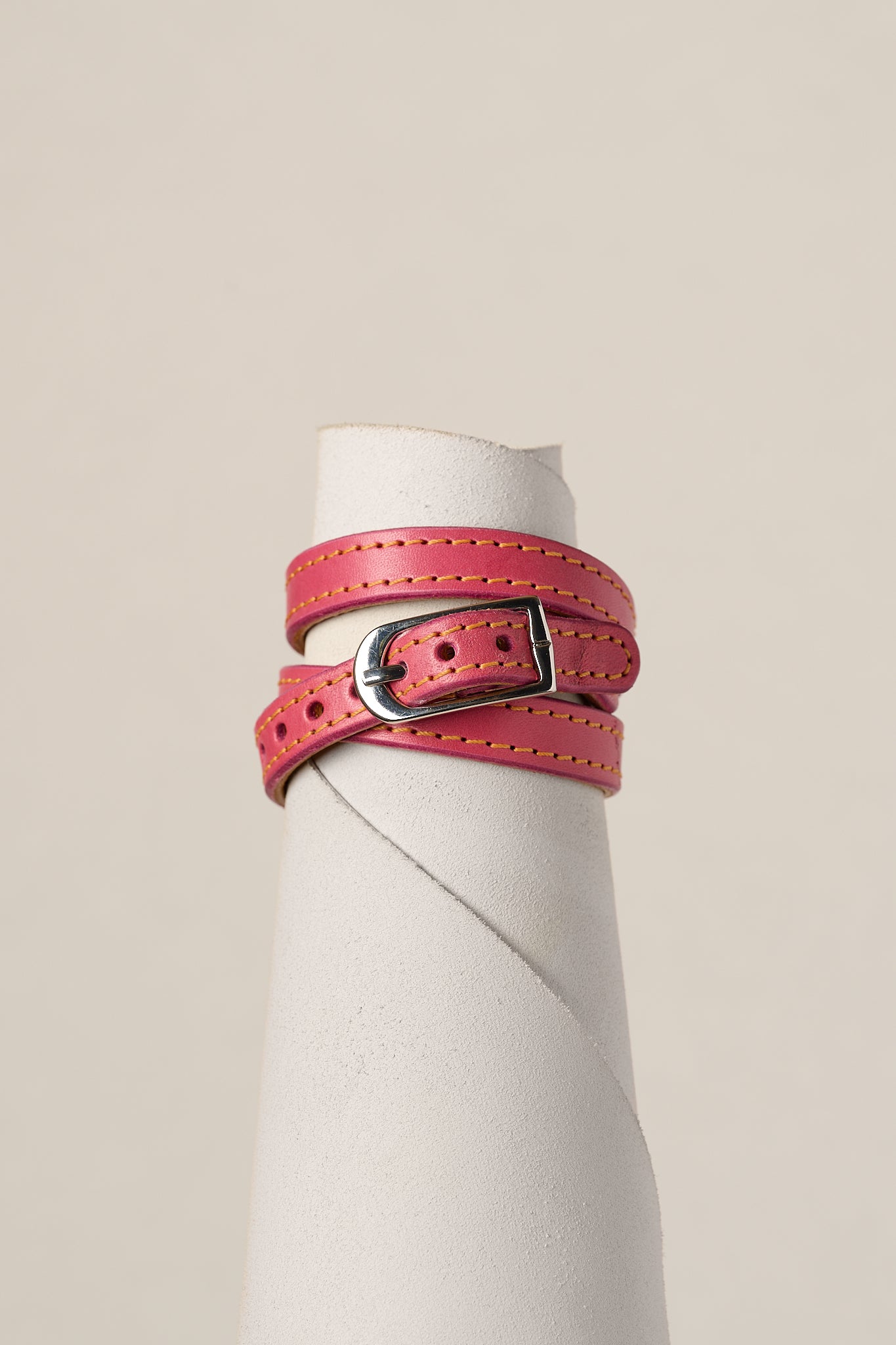A pink leather belt with a silver-toned metal buckle, wrapped around a white cylindrical display stand against a light beige background.