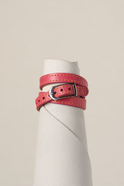 A pink leather belt with a silver-toned metal buckle, wrapped around a white cylindrical display stand against a light beige background.