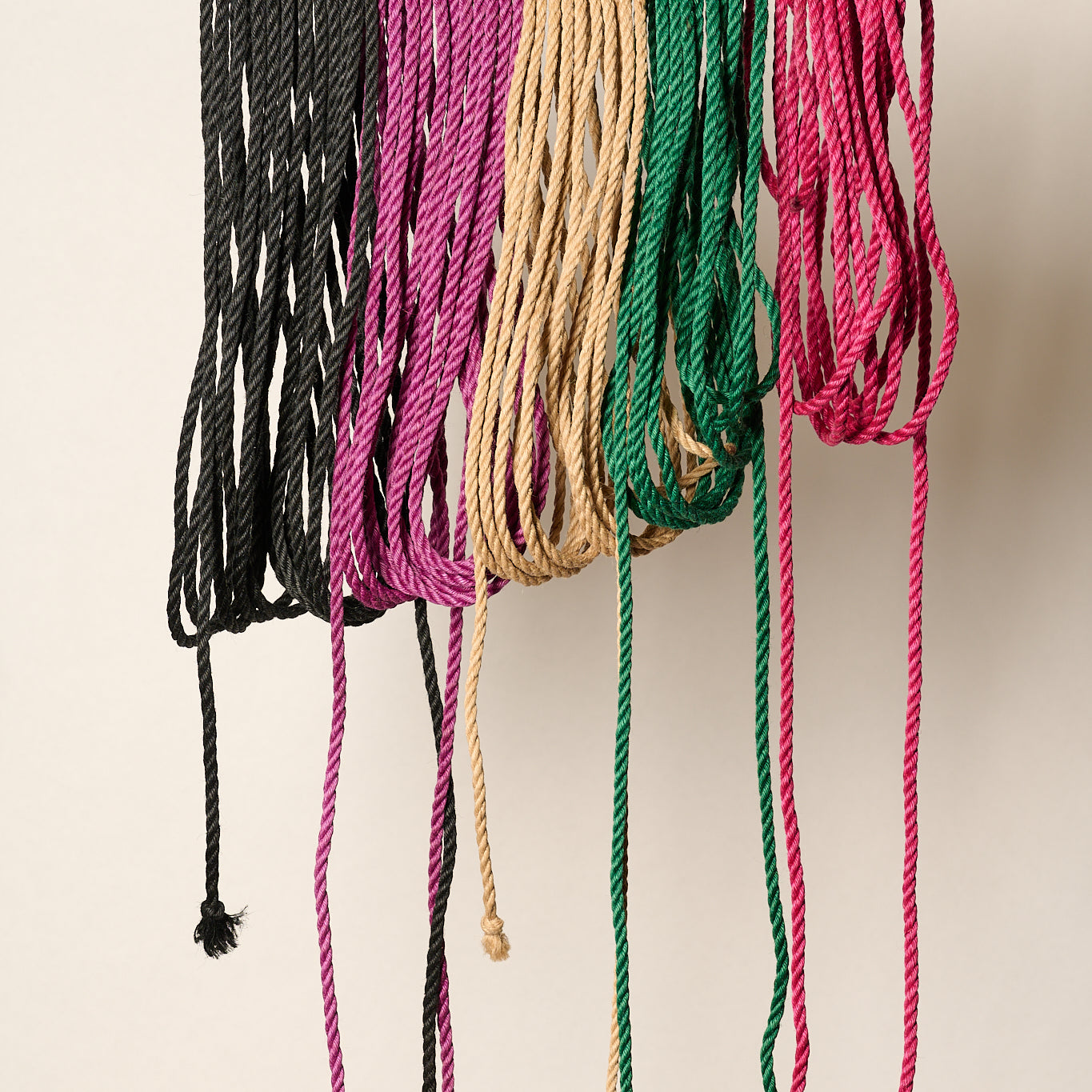 Six braided jute rope cords in black, magenta, tan, green, and pink hanging from a metal rod against a plain off-white background.