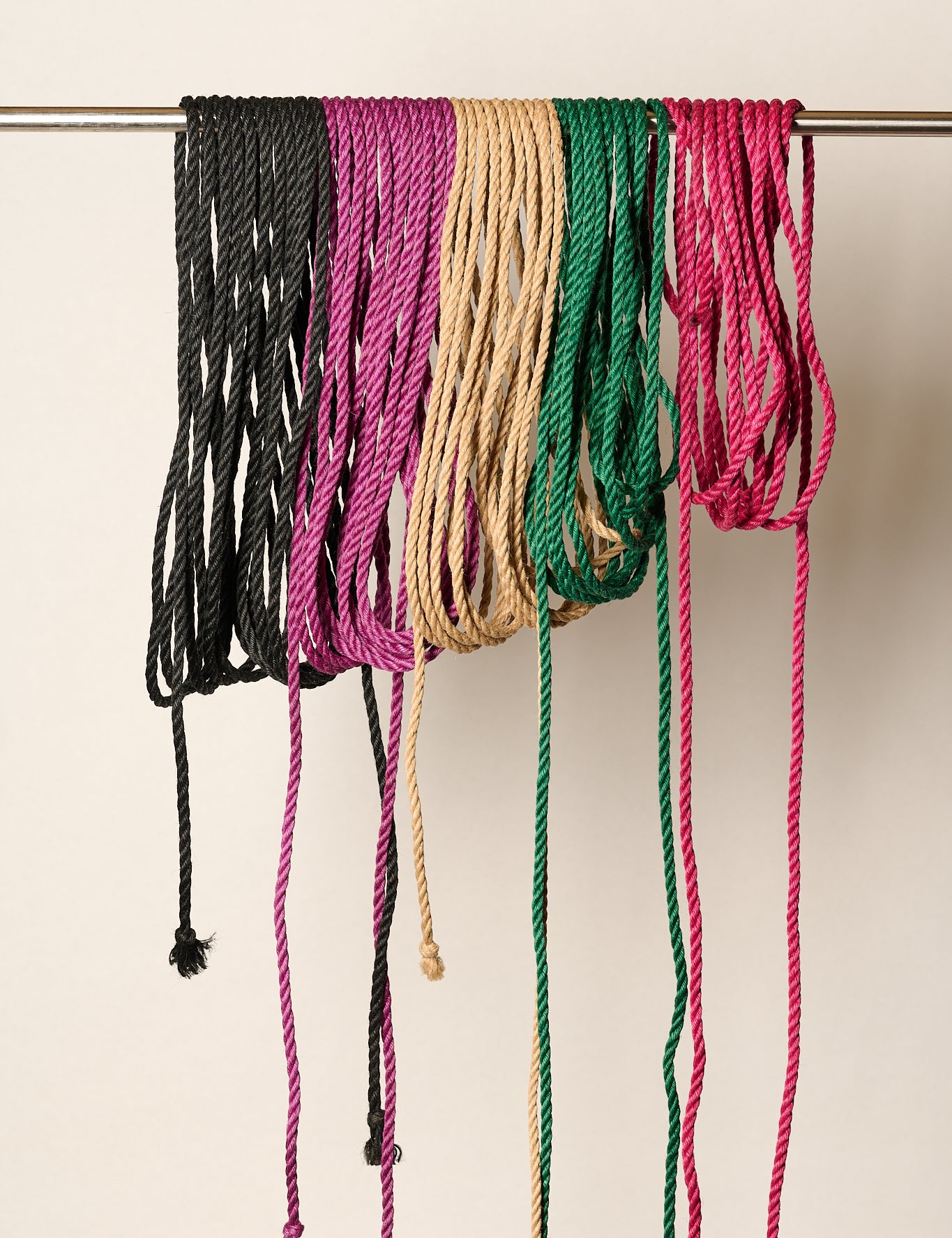 Six braided jute rope cords in black, magenta, tan, green, and pink hanging from a metal rod against a plain off-white background.