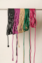 Six braided jute rope cords in black, magenta, tan, green, and pink hanging from a metal rod against a plain off-white background.