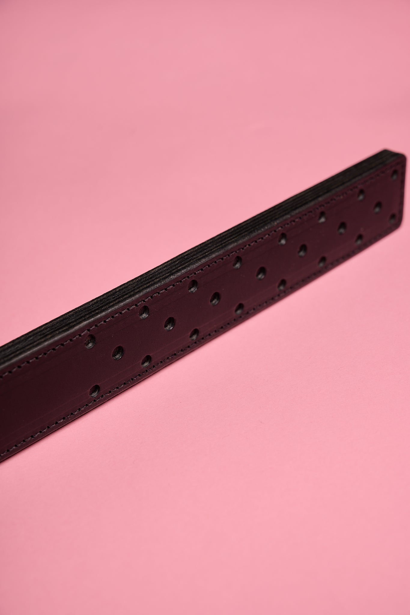 A black leather strap with a fenestrated surface, laid on a pink background.