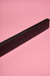 A black leather strap with a fenestrated surface, laid on a pink background.