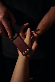 Lovey Subby burgundy/pomegranate leather portable pocket paddle with brass rivets and black thread in a mans hand laying on a womans hand.