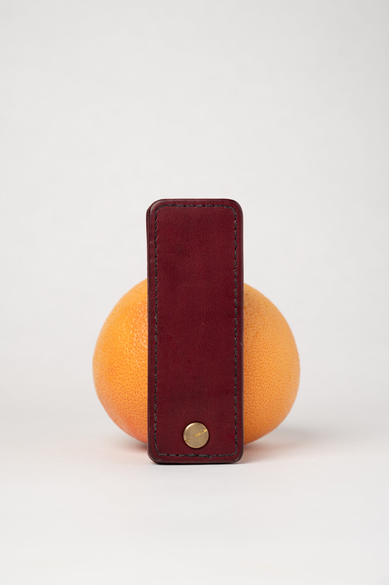 foldable burgundy leather travel size belt with rivets on white background with grapefruit behind.