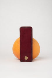 foldable burgundy leather travel size belt with rivets on white background with grapefruit behind.