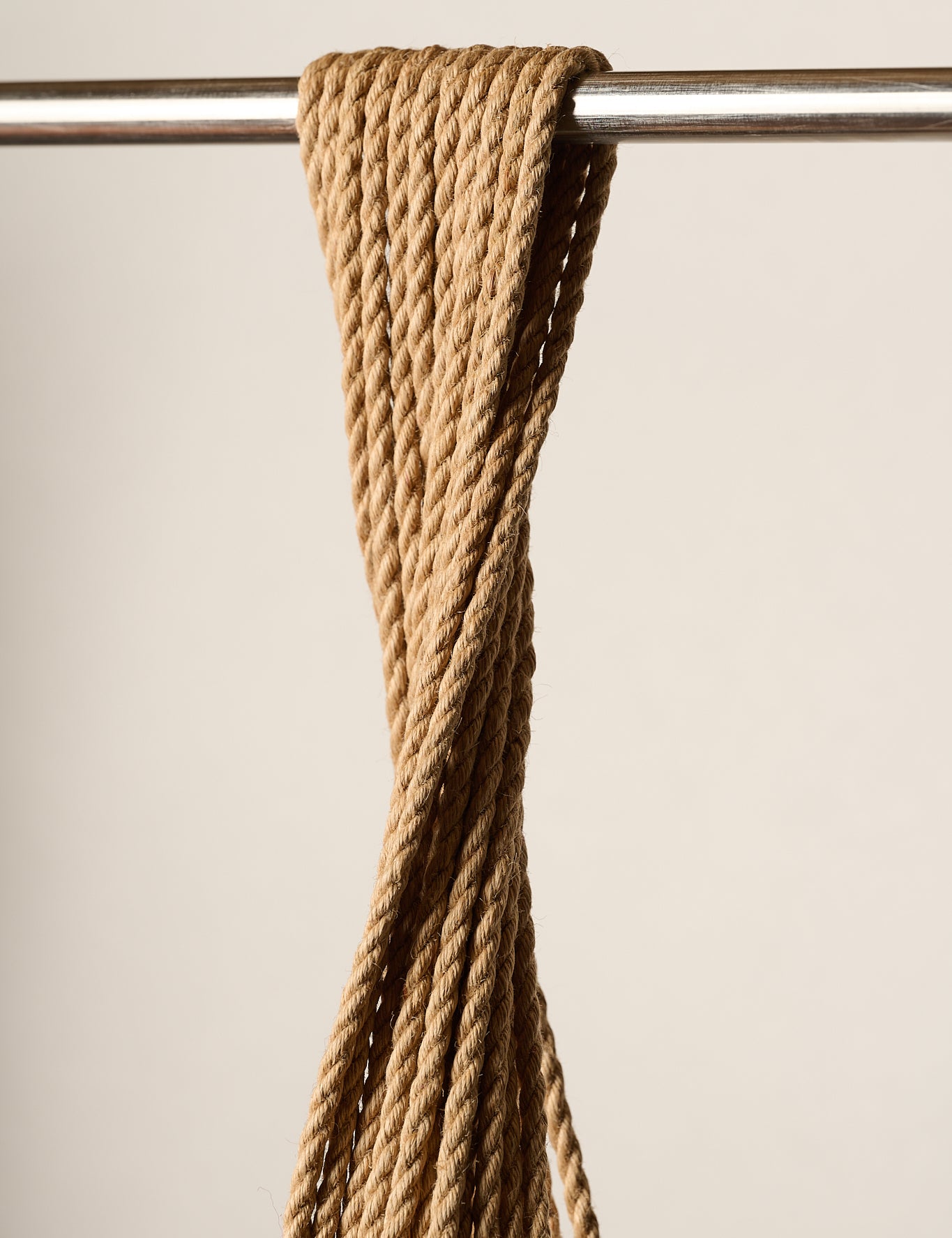A close-up of a 6mm, twisted tan jute, rope hanging from a horizontal metal rod against a plain beige background.