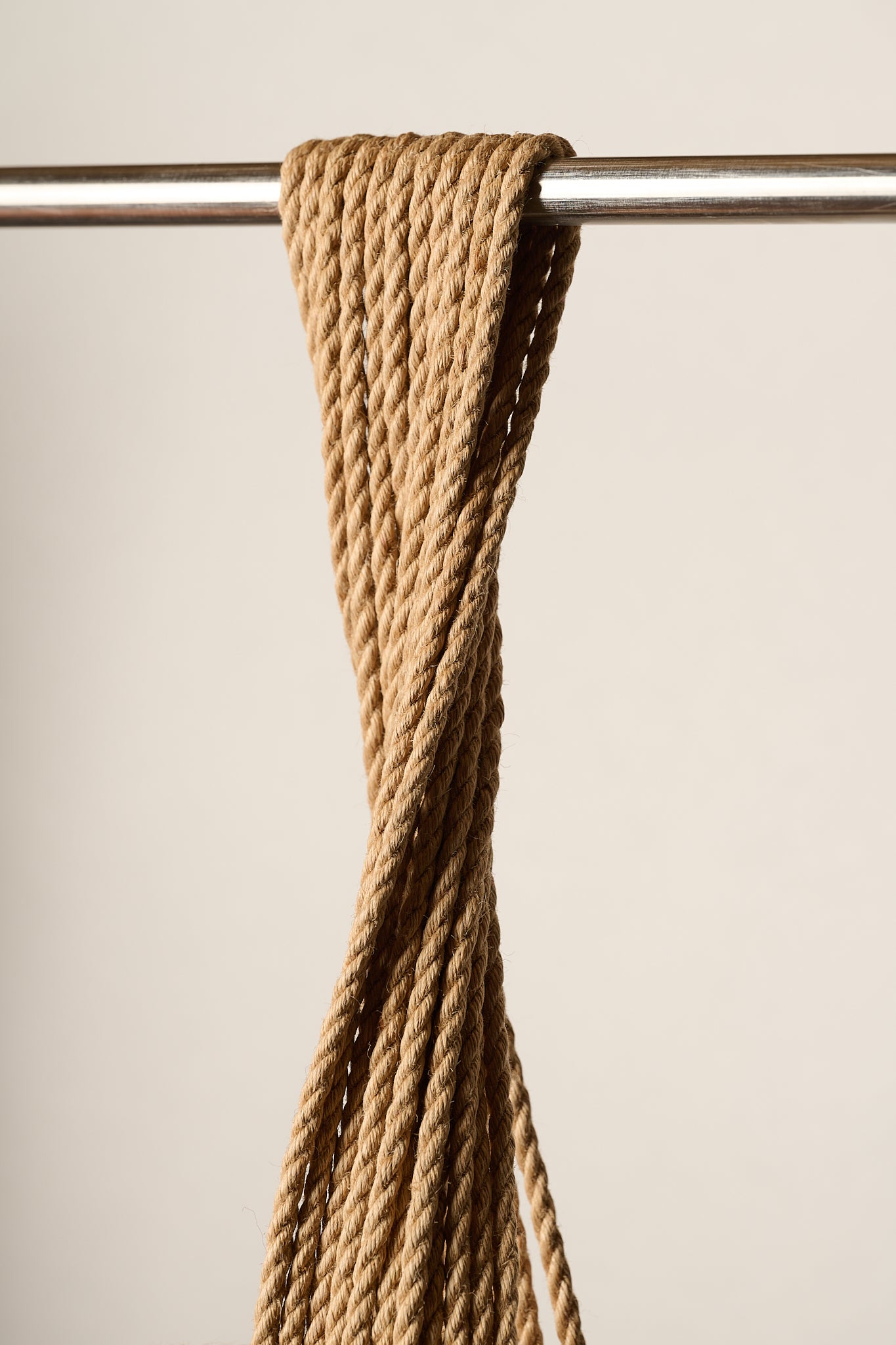 A close-up of a 6mm, twisted tan jute, rope hanging from a horizontal metal rod against a plain beige background.