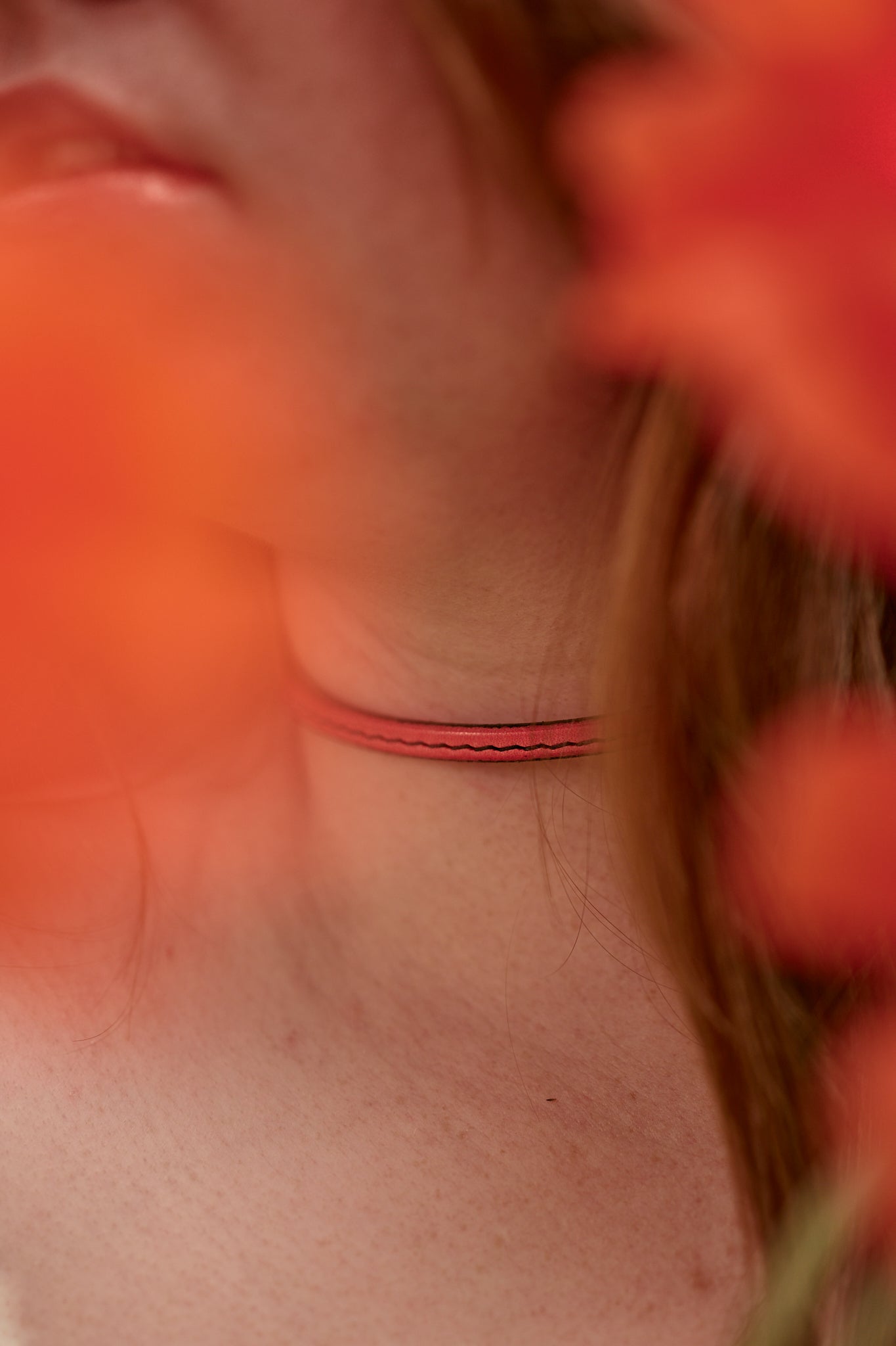 Close-up of a person's neck with a thin red necklace, surrounded by blurred orange flowers.