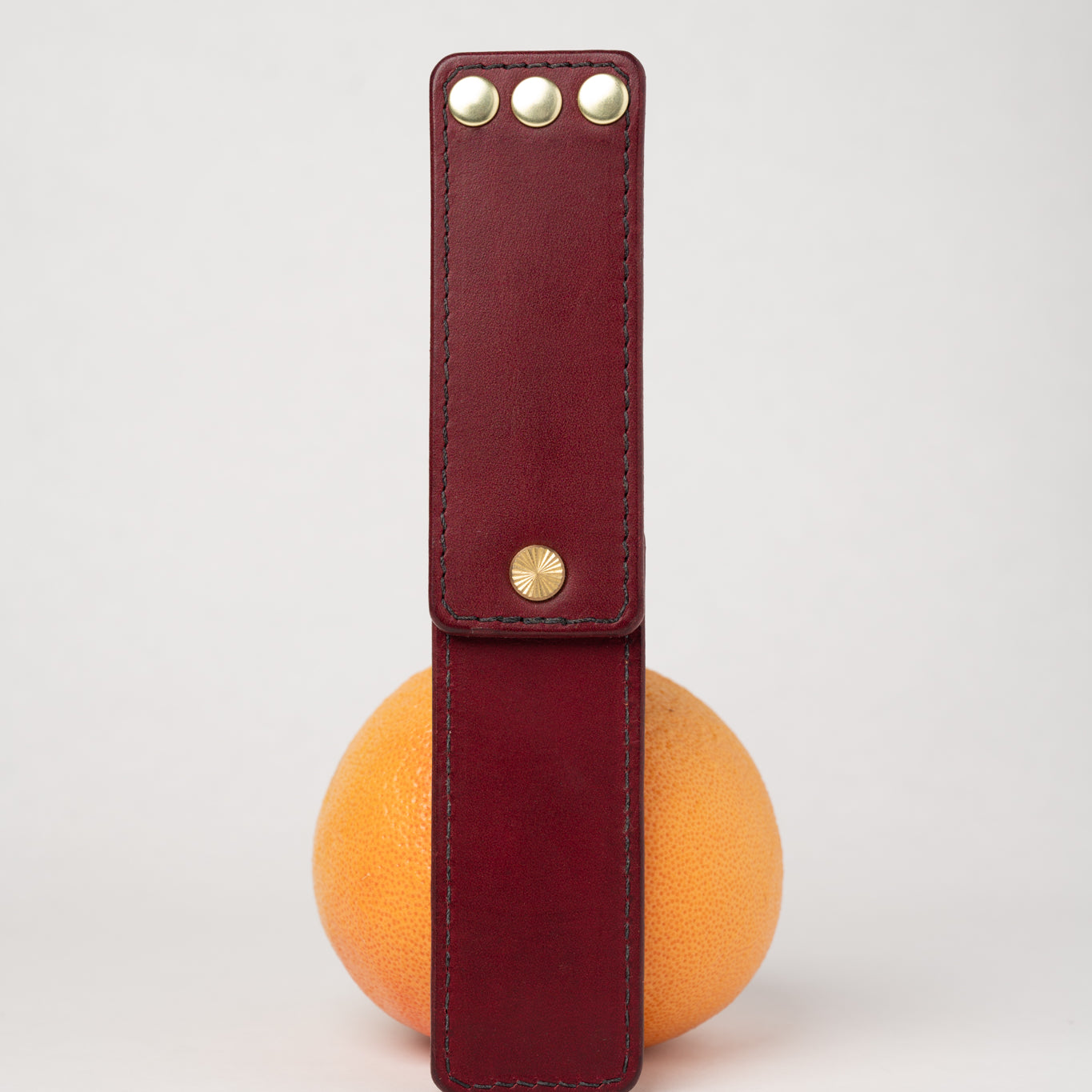foldable burgundy leather travel size belt with rivets on white background with grapefruit behind.