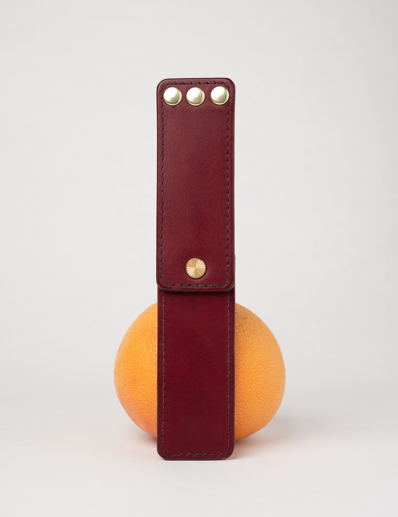 foldable burgundy leather travel size belt with rivets on white background with grapefruit behind.