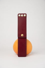 foldable burgundy leather travel size belt with rivets on white background with grapefruit behind.
