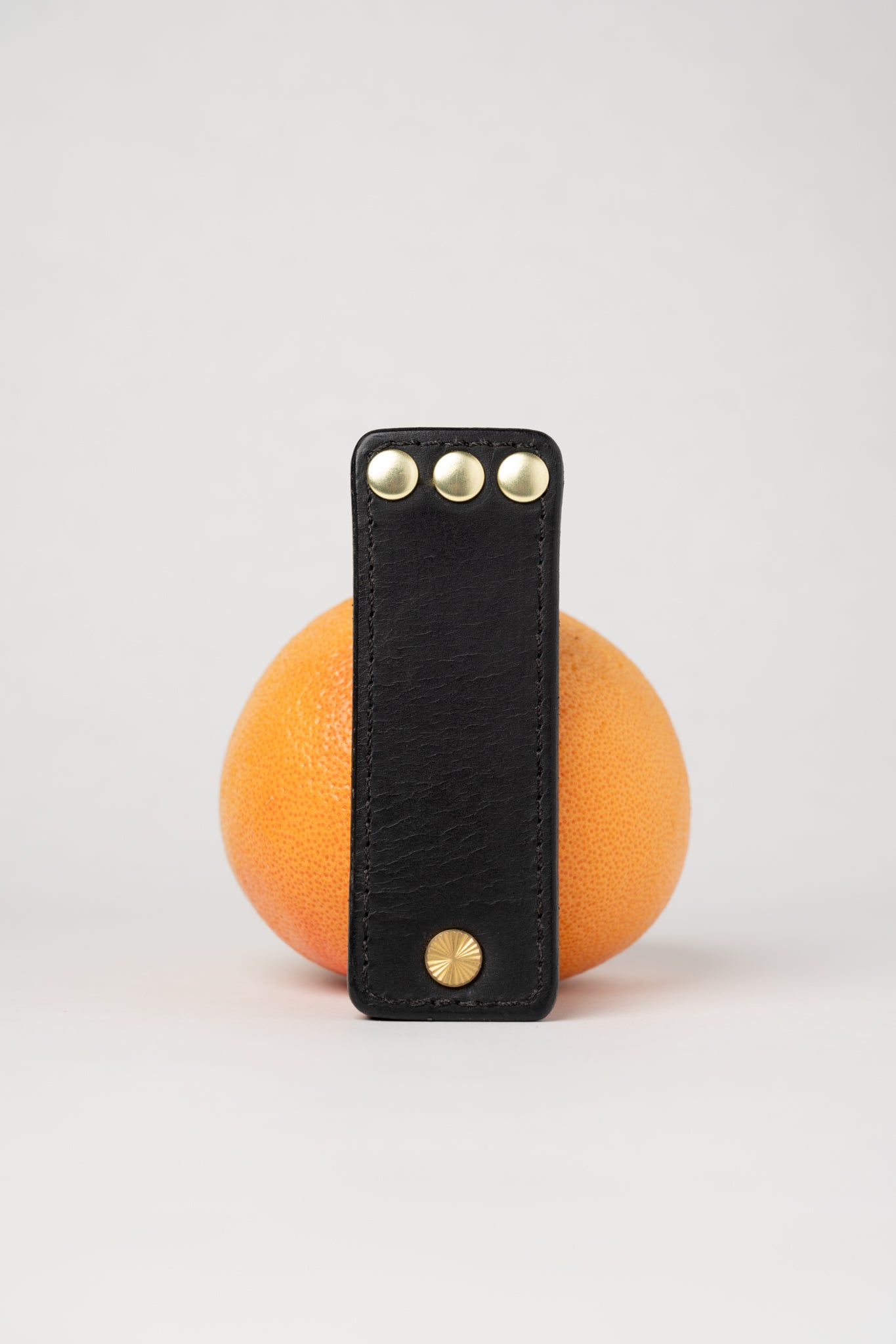 foldable black  leather travel size belt with rivets on white background with grapefruit behind.