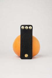 foldable black  leather travel size belt with rivets on white background with grapefruit behind.